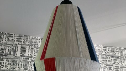Ein hängender Lampenschirm, mit rotem, blauem und weißen Garn bespannt, schwebt vor einer grauen, grafischen Tapete.