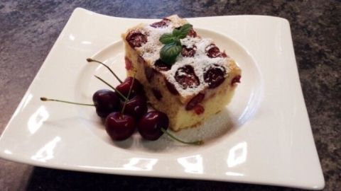 Ein Kirschkuchen liegt auf einem weißen Teller, dekoriert mit Kirschen und einem Hauch von Minze, in einer modernen Küche.