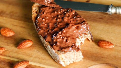 Ein Stück Brot ist mit Nusscreme bestrichen und auf einem Holzbrett platziert, umgeben von einigen Mandeln.