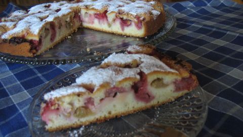 Eine Rhabarber-Erdbeer-Joghurt-Torte liegt auf einem dekorativen Glas-Teller. Zarte Stücke sind angeschnitten und mit Puderzucker bestäubt, auf einem karierten Tischcloth.