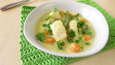 Eine Schüssel mit einfacher Fischsuppe, die mit buntem Gemüse und frischen Kräutern auf einem grünen Platz serviert wird.