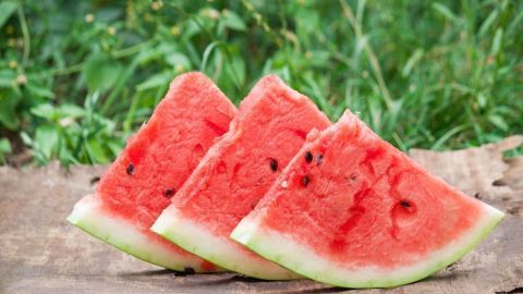 Drei Wassermelonenstücke liegen auf einer Holzoberfläche, umgeben von grünem Gras im Freien, bereit zum Genießen.