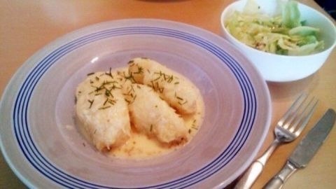 Auf einem Teller sind drei große, in Scheiben geschnittene Kartoffelklöße zu sehen, die mit Kräutern bestreut sind. Daneben steht eine Schüssel mit frischem Salat.