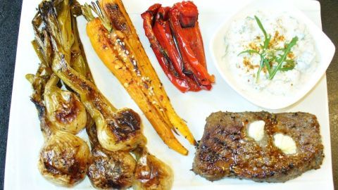 Geschmorte scharfe Frühlingszwiebeln liegen neben einem Steak, Karotten und einer Sauce auf einem weißen Teller.