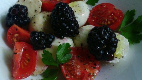 Ein bunter Salat aus Mozzarella, frischen Brombeeren und Tomaten liegt in einer schlichten weißen Schüssel, umgeben von Petersilie.