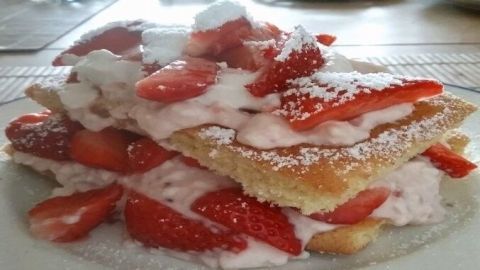 Ein schichtiger Erdbeerkuchen liegt auf einem Teller, bedeckt mit frischen Erdbeeren und Sahne, garniert mit Puderzucker.