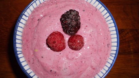 Ein Schüsselchen mit Frischkornbrei, cremig und rosa, garniert mit drei frischen Beeren in einem rustikalen Holzumfeld.