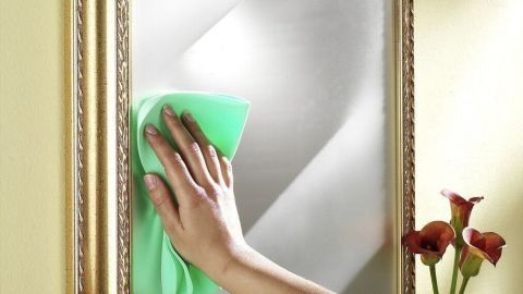 Eine Hand mit einem grünen Tuch reinigt einen Spiegel, um eine klare Sicht in einem hellen Raum zu gewährleisten.