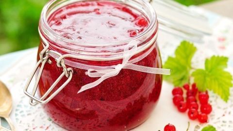 Ein Glas Wassermelonen-Johannisbeer-Gelee steht auf einem hübsch dekorierten Tisch, umgeben von grüner Wiese und roten Johannisbeeren.