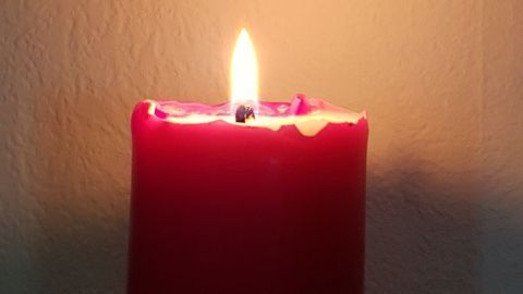 Eine rote Kerze leuchtet hell auf einem Tisch, während im Hintergrund ein Bild von kahlen Bäumen an der Wand hängt.