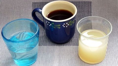 Drei Gläser stehen auf einem Tisch: ein blaues Glas mit Wasser, eine Tasse Kaffee und ein weißer Saft.
