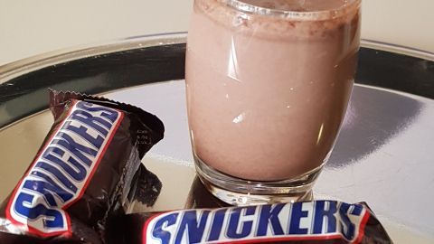 Ein Glas Snickers-Likör steht auf einem silbernen Tablett, umgeben von Snickers-Riegeln, vor einem modernen Hintergrund.