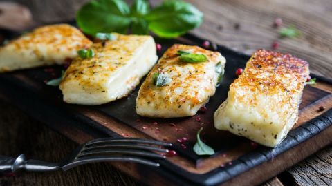 Gegrillter Halloumi liegt auf einem Holzbrett, goldbraun und knusprig, umgeben von frischen Kräutern und einer Gabel.