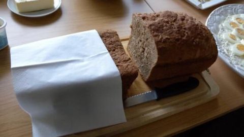 Frisches 3-Minuten-Brot liegt auf einem Holzbrett, während ein Stück abgeschnitten ist; die Umgebung zeigt eine gedeckte Tafel.