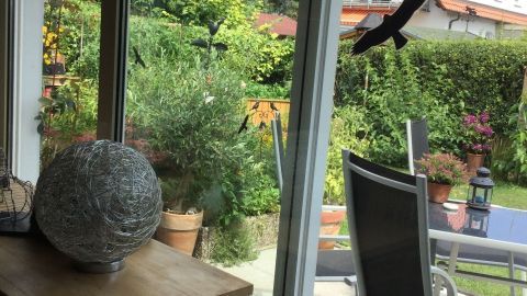 Ein großer Balkon mit einem Tisch und Stühlen bietet einen Blick auf den grünen Garten, während große Fensterflächen Licht einlassen.