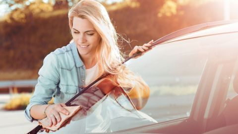 Eine Frau reinigt mit einem Tuch die Windschutzscheibe eines Autos in einer sonnigen Umgebung.