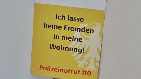 Eine gelbe Türanhängerkarte hängt an einer Wohnungstür, die sagt: "Ich lasse keine Fremden in meine Wohnung!" und verweist auf Polizei.