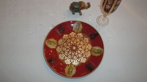Ein dekorierter Glasteller mit goldenen Mustern liegt auf einem weißen Tisch, umgeben von einem kleinen Elefanten und einem Glas.