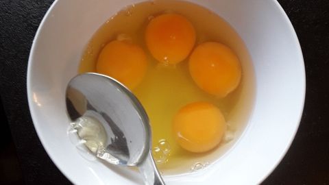 In einer weißen Schüssel liegen vier rohe Eier, während ein Löffel im flüssigen Eigelb schwenkt, um sie zu vermischen.