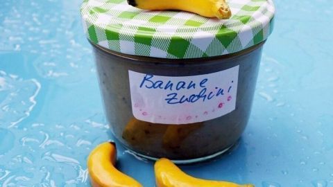 Ein Glas Marmelade mit einem grünen karierten Deckel steht auf einer blauen Oberfläche, neben zwei kleinen Bananen.