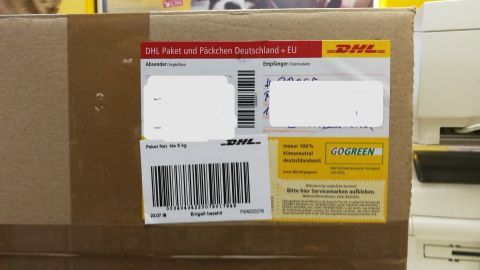 Ein braunes Paket mit DHL-Etikett liegt auf einem Tisch in einem Postbüro, umgeben von gelben Wänden und Druckern.