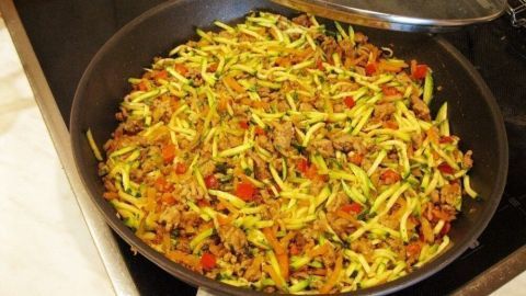 Eine große Pfanne enthält eine bunte Mischung aus Zucchini und Hackfleisch, die auf einem Herd brutzelt.