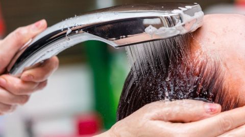 Ein Wasserstrahl aus einer Dusche reinigt gefärbte Haare, während eine Hand den Kopf sanft hält.