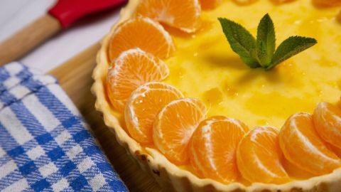 Ein bunten Schüttelkuchen mit Mandarinen und Minze dekoriert, liegt auf einem Holzbrett neben einem karierten Geschirrtuch.