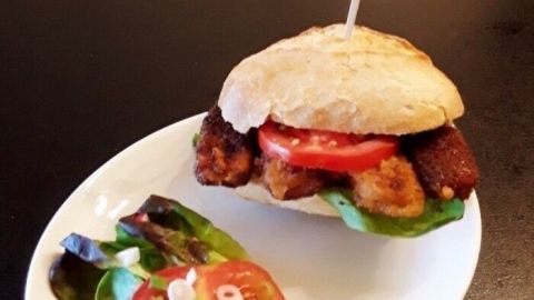 Ein Fischstäbchenburger liegt auf einem weißen Teller, garniert mit Salat und Tomaten, auf einem dunklen Tisch.