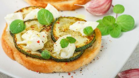Zucchinitoast liegt appetitlich auf einem Teller, belegt mit Zucchinischeiben und frischem Kräutern, umgeben von Knoblauchzehen.