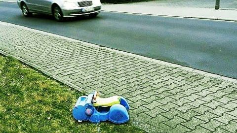 Ein blaues Kinderspielzeug liegt auf dem Gehweg, während ein Auto vorbeifährt, was das Risiko von Rasern verdeutlicht.