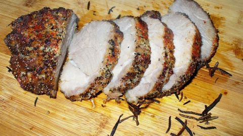 Schweinelachs liegt auf einem Holzbrett, in saftige Scheiben geschnitten, mit bunter Pfefferkruste und Kräutern garniert.