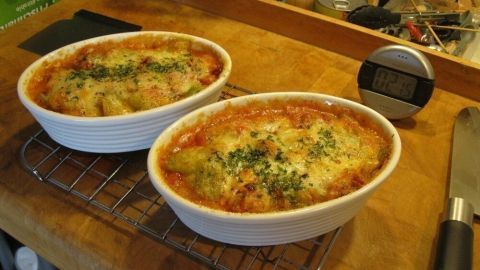 Zwei heiße Fisolenaufläufe mit Chorizo und Emmentaler stehen auf einem Holzbrett, umgeben von Küchengeräten.