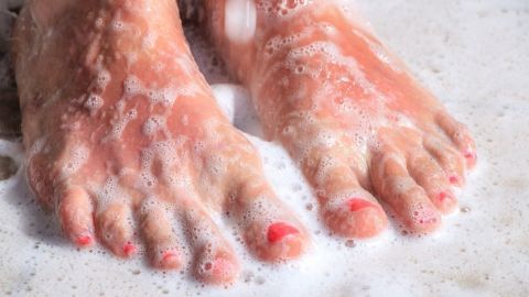 Zwei Füße mit rotem Nagellack stehen im seifigen Wasser, während Schaum sie umspült und eine sanfte Reinigung symbolisiert.