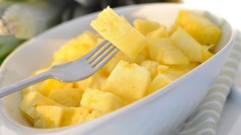Saure Ananasstücke liegen in einer weißen Schüssel, während eine Gabel sie aufnimmt. Im Hintergrund steht eine frische Ananas.