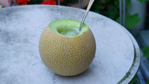 Eine aufgeschnittene Melone mit einem Löffel steht auf einem Tisch, umgeben von blühenden Pflanzen im Hintergrund.