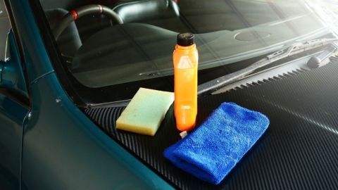 Eine Flasche Autopolitur steht neben einem gelben Schwamm und einem blauen Mikrofasertuch auf einer Autoscheibe in einer Garage.