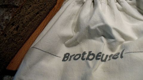 Ein Baumwollsack mit der Aufschrift „Brotbeutel“ liegt neben einem Holzbrett, das frisches Brot enthält.