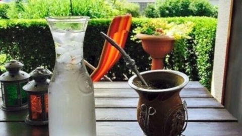Ein gefüllter Krug mit Wasser steht neben einem traditionellen, kunstvoll gestalteten Mategefäß auf einem Tisch im Freien, umgeben von grünem Garten.