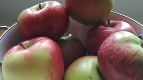 Frische Äpfel liegen übereinander in einer Schüssel, umgeben von farbigen Pflanzenkübeln im Hintergrund.