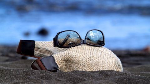 Eine Sonnenbrille liegt auf einem Strohhut, der im Sand am Strand platziert ist, während das Wasser im Hintergrund glitzert.