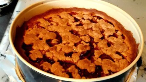 Ein runder Kuchen, der frisch gebacken wurde, steht in einer Backform auf einer Küchenarbeitsplatte und zeigt eine goldene Mürbeteig-Oberfläche.