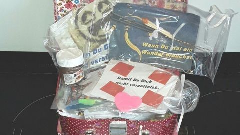 Ein bunter Koffer steht offen auf einer schwarzen Oberfläche, gefüllt mit verschiedenen Geschenken und buntem Papier.