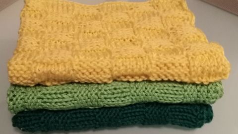 Gestapelte, handgestrickte Spültücher in Gelb, Grün und Dunkelgrün liegen auf einer glatten Oberfläche.