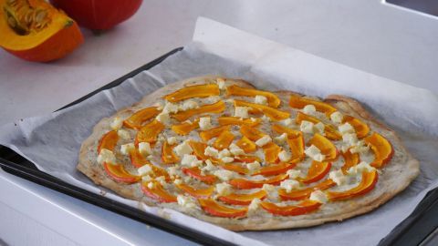 Ein Flammkuchen mit Kürbis und Feta liegt auf einem Backblech, umgeben von frischem Kürbis, bereit zum Backen.