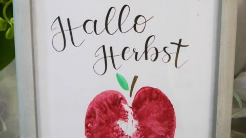 Ein gerahmtes Apfelbild mit dem Text "Hallo Herbst" steht auf einem Tisch, umgeben von grünen Pflanzen in einem hellen Raum.