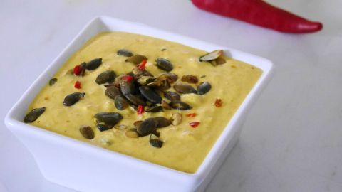 Ein herzhafter Kürbis-Aufstrich in einer Schüssel, garniert mit Kürbiskernen, steht neben frischen Brötchen und einer Chili.