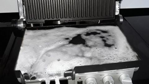 Ein Grillgerät steht über einer offenen Geschirrspülmaschine und ist voller Seife. Küchenelemente sind darin zu sehen.