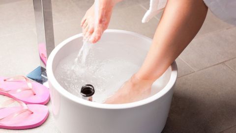 Zwei Füße tauchen in ein Wasserbecken, um kalte Füße zu wärmen, während rosa Hausschuhe daneben liegen.