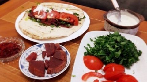 Auf einem Tisch liegt ein Falafel-Wrap, während frische Tomaten, grüner Salat und Soßen zur Zubereitung bereitstehen.